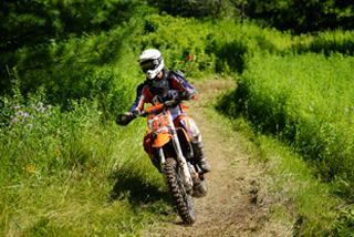 A person in protective gear and a white helmet rides an orange dirt bike along a narrow dirt trail through lush green forest and tall grass