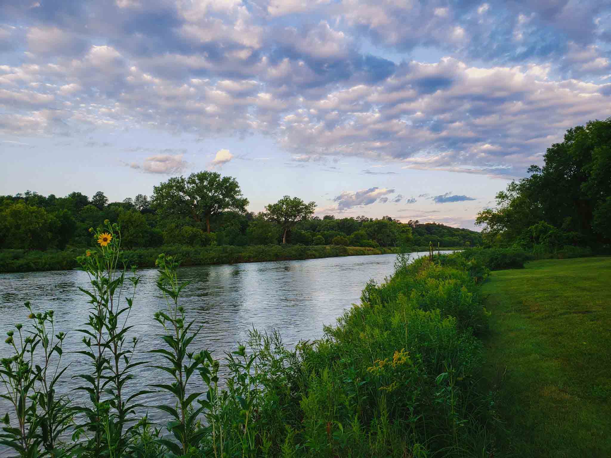 A wide river flows through a lush green landscape under a vast, cloudy sky at dusk