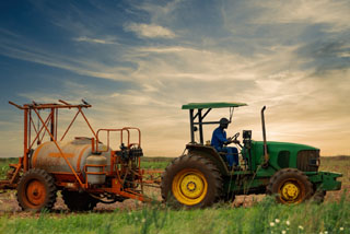 A farmer operates a green tractor pulling a large orange tank sprayer across a field under a vast, clouded sky at dusk