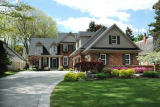 A large, two-story suburban house with a brick and siding exterior, multiple gables, and dark shutters, set behind a manicured lawn with colorful landscaping and a wide concrete driveway