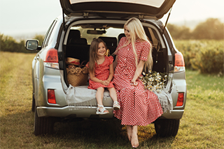 A mother and young daughter in matching red dresses sit smiling in the open trunk of a silver SUV parked in a grassy field